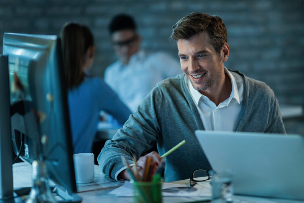Happy male entrepreneur using desktop PC while working in the office. There are people in the background.